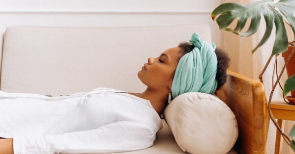 A woman in a head scarf relaxes on a beige sofa indoors surrounded by plants, peaceful scene.